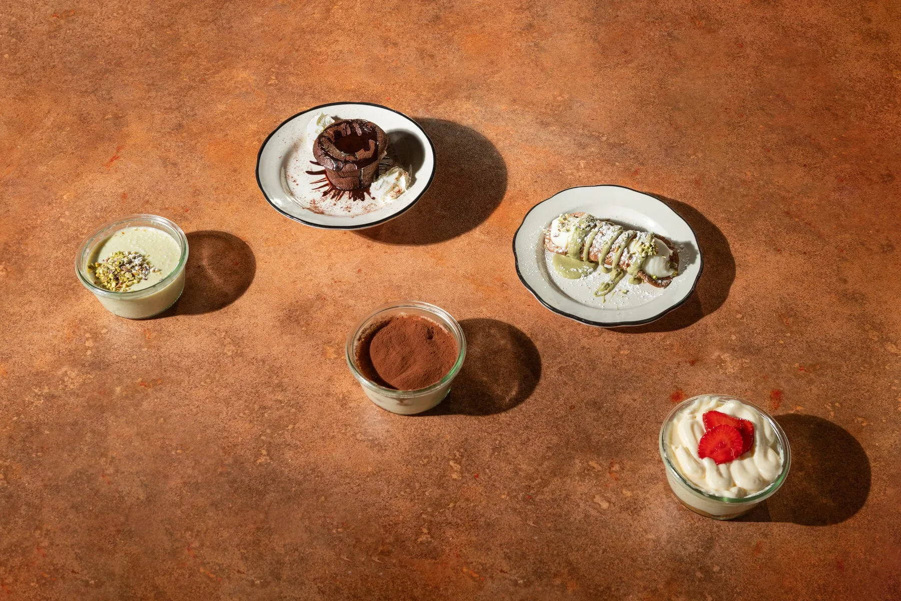 Assortiment de desserts vu du dessus : panna cotta à la pistache, moelleux au chocolat, tiramisu, cannolo à la pistache et tiramisu à la fraise