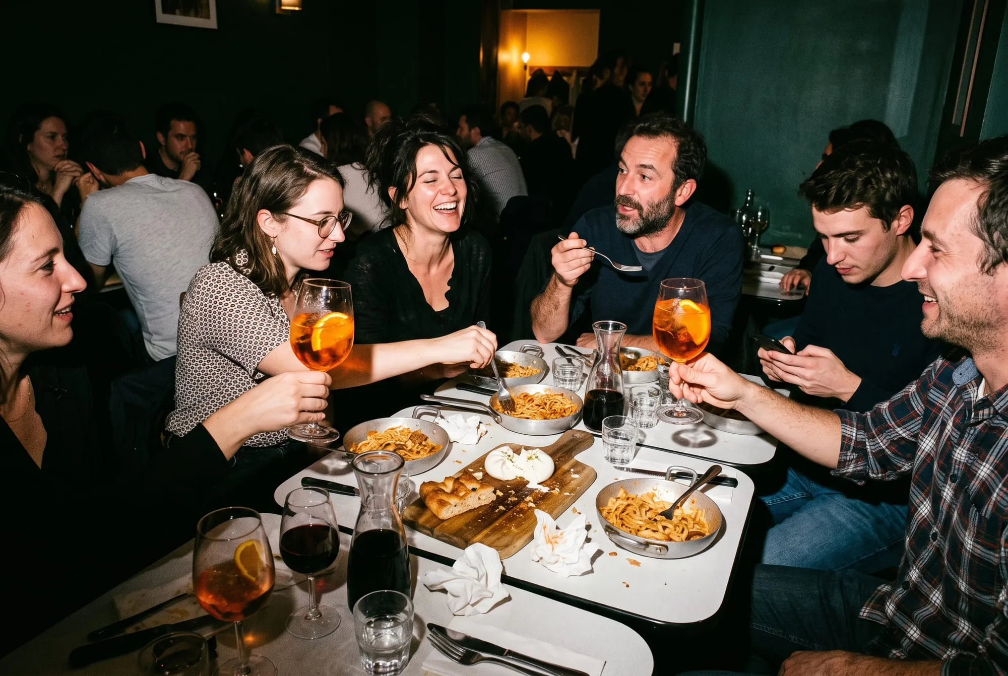 Une bande d’amis rit et bavarde autour de tables rapprochées, le soir, deux spritz levés : pâtes servies au poêlon, burrata ouverte sur une planche de focaccia, carafes de vin rouge
