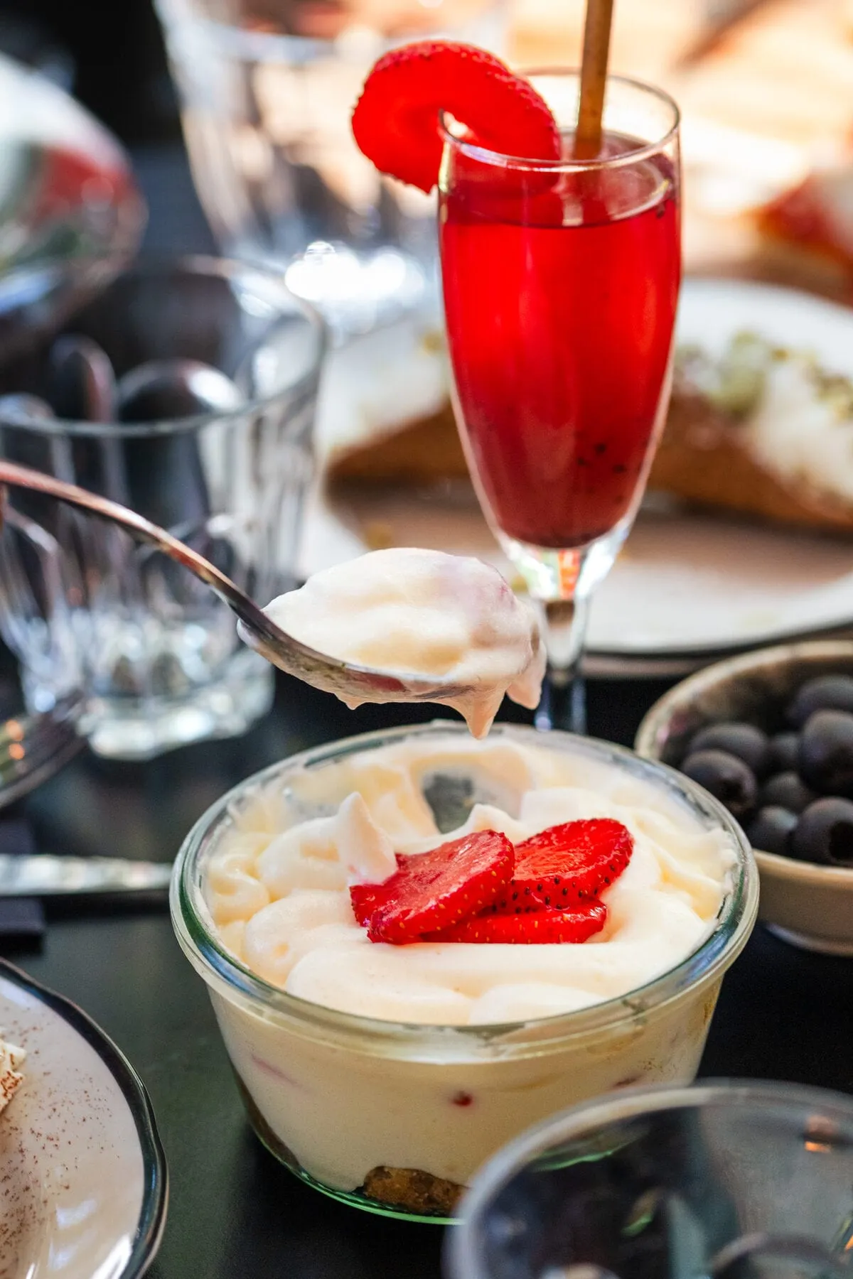Tiramisu à la fraise en pot de verre, cuillère levée, et cocktail rouge à la fraise en arrière-plan