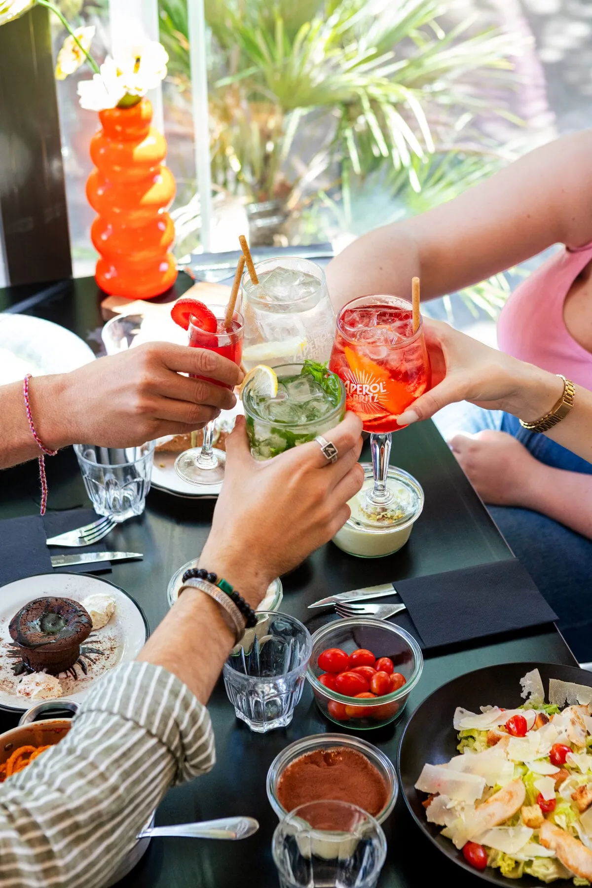 Quatre convives trinquent avec des spritz et des mojitos autour d’une table garnie, en terrasse